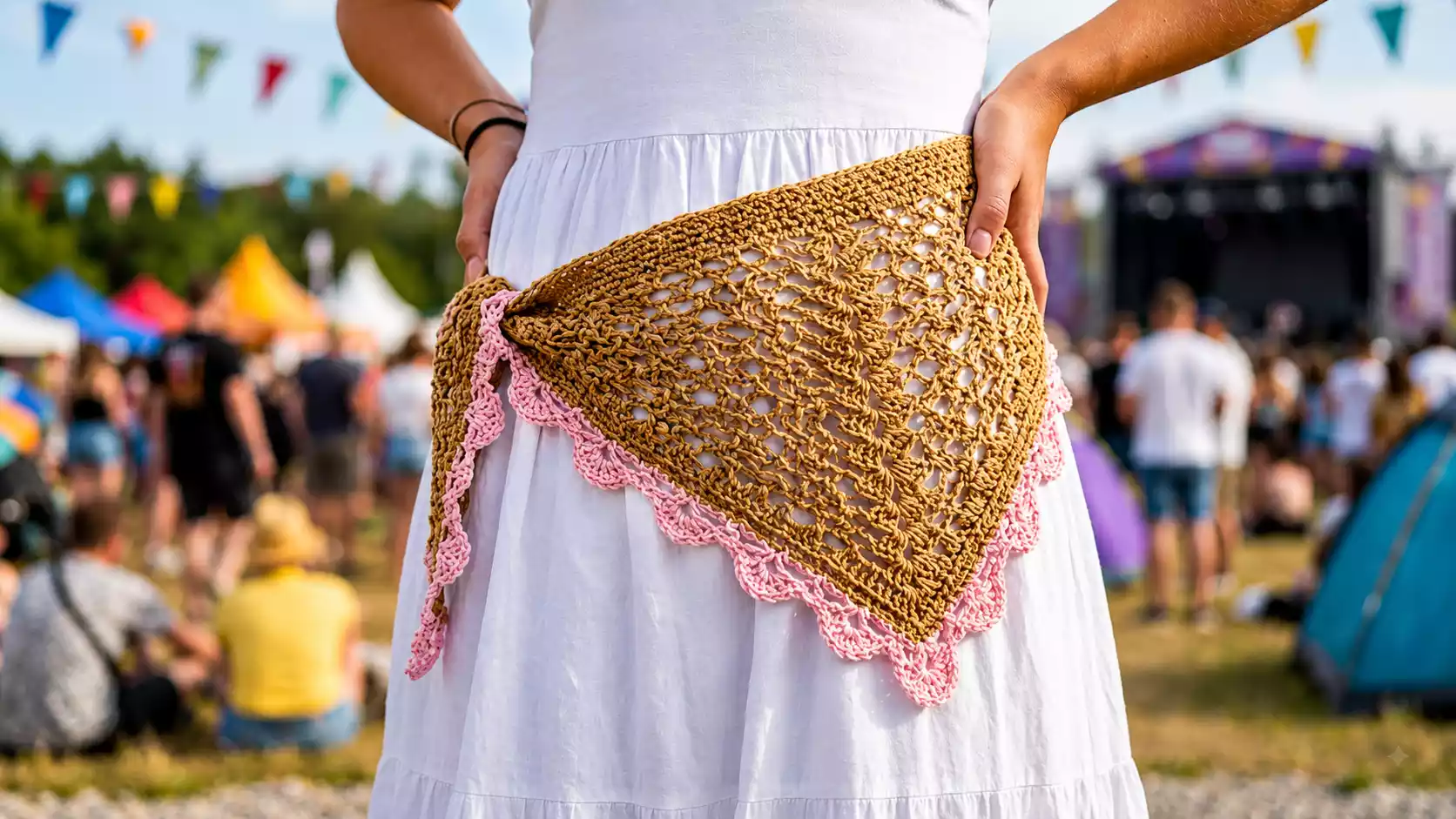 Mini chal o bandana Luxia tejida a crochet en tono camel con borde rosa, de diseño calado triangular, anudada sobre un vestido blanco durante un festival al aire libre con ambiente alegre y veraniego.