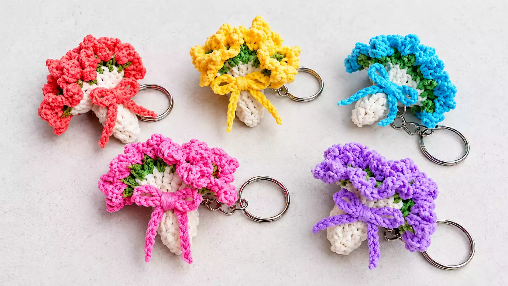 Cinco llaveros de pequeños ramos de flores tejidos a crochet en colores rosa, coral, amarillo, azul y lila, con envoltura blanca, hojas verdes, lazos decorativos y argollas metálicas.