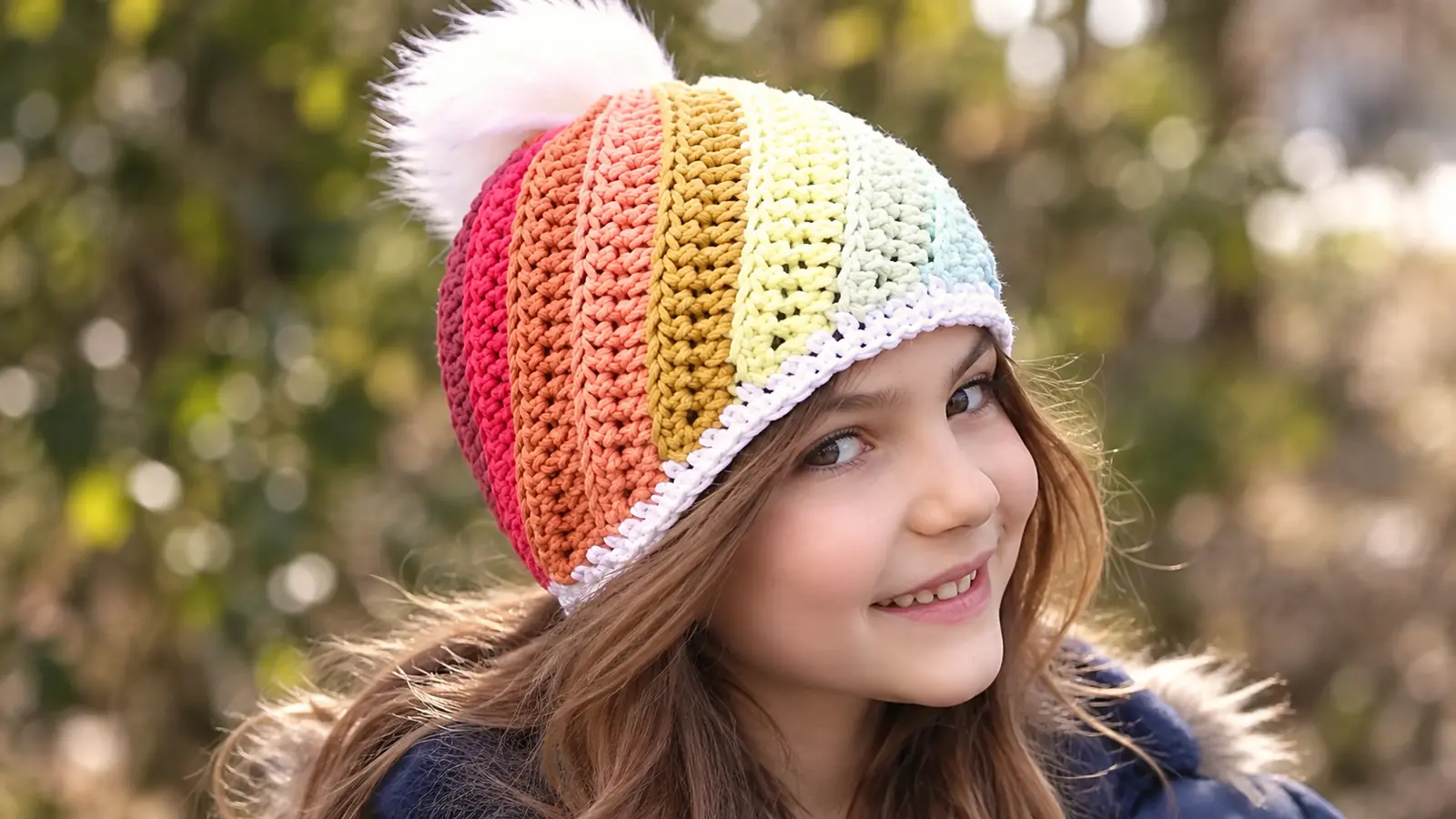 Preadolescente luciendo un gorro Lollypop tejido a crochet, con franjas multicolor, punto texturizado y pompón blanco.