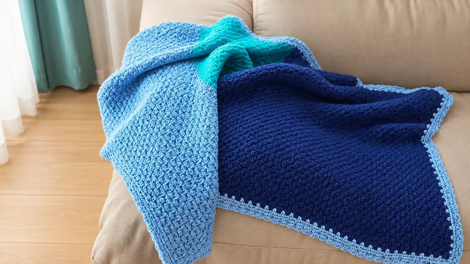 Manta tejida a crochet en tonos azul y turquesa, con borde celeste decorativo, sobre un sofá beige.