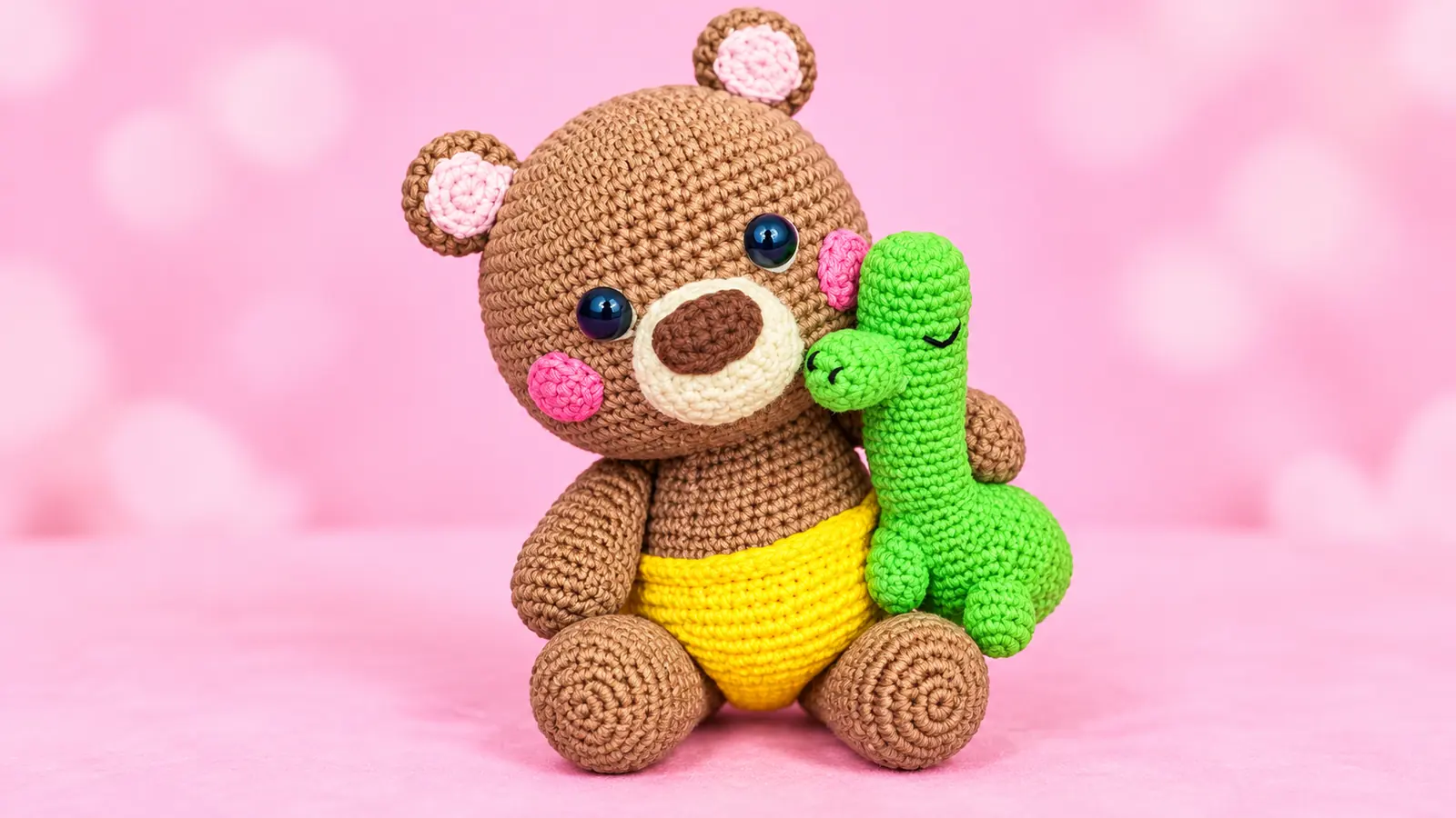 Osito amigurumi tejido a crochet en color café, con pañal amarillo y mejillas rosadas, abrazando un dinosaurio verde sobre fondo rosa.