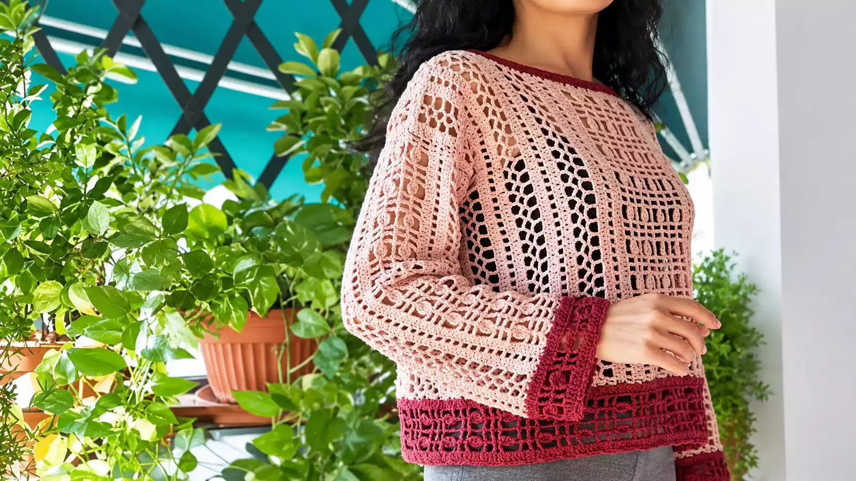 Mujer luciendo el jersey Dafne tejido a crochet en rosa claro, con motivos geométricos calados, mangas largas y amplias y bordes en tono frambuesa, junto a plantas en una terraza.